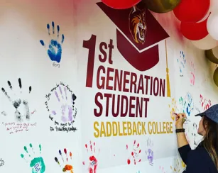 A wall with the words 1st Generation Student Saddleback College is decorated with the painted handprints of students. A female student is pictured writing her name on her handprint.